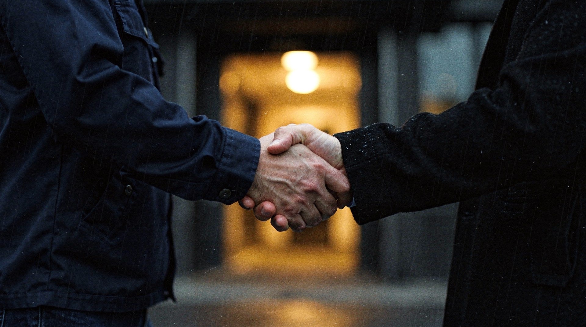 Handschlag zweier Männer in dunklen Jacken im Regen vor einem beleuchteten Hauseingang