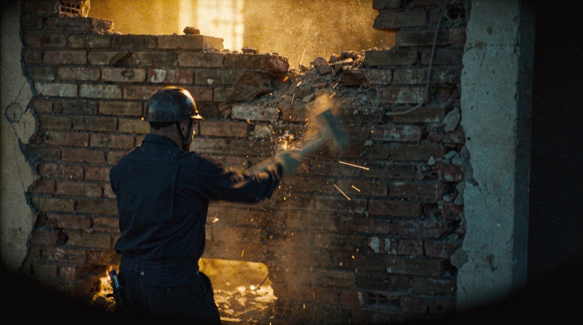 Arbeiter mit Helm schlägt mit dem Hammer eine Ziegelwand auf, Funken und Staub im Gegenlicht